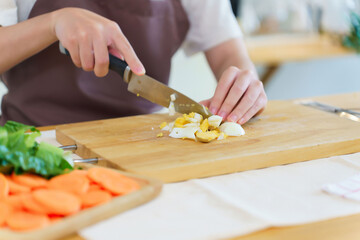 Closeup hands of asian women chopping boil egg with knife on cutting board and slice fresh vegetable to preparing ingredients for cooking breakfast meal while making healthy food lifestyle in kitchen