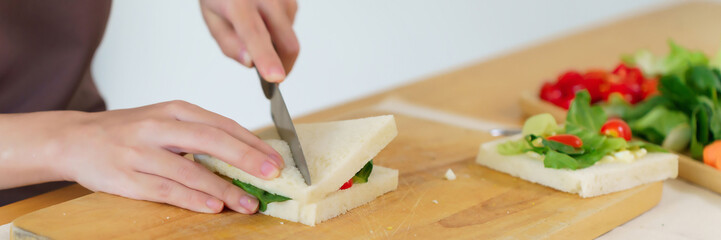 Asian women making delicious sandwich with vegetables and mayonnaise to cutting sandwich with knife on cutting board while cooking breakfast meal and making healthy food lifestyle in kitchen at home