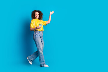 Full body photo of attractive young woman walk wave hand hold device dressed striped yellow clothes isolated on blue color background