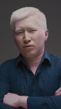 Portrait of an Asian albino guy. Eastern man with a genetic mutation of pigment, white hair. The arms are crossed on the chest. The male is wearing a dark shirt in the studio. Abnormal deviations