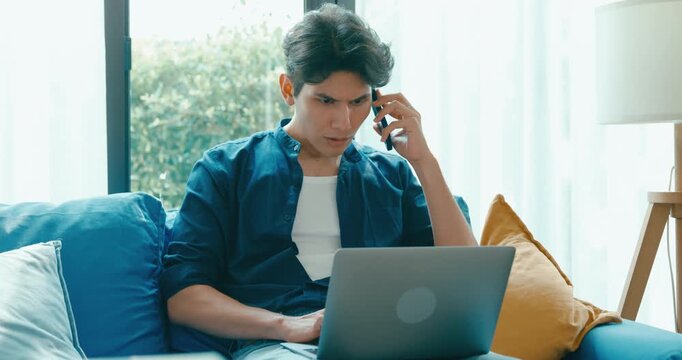 Positive young freelance Asia male use laptop and talk on cell phone remote working on sofa in living room at home office. Work from house, financial plan, remotely digital nomad job and Gen Z talent.