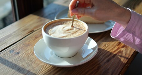 Woman stirs decorative foamy coffee drink at breakfast in coffeeshop closeup. Female takes spoon with dripping froth out of beverage at wooden table