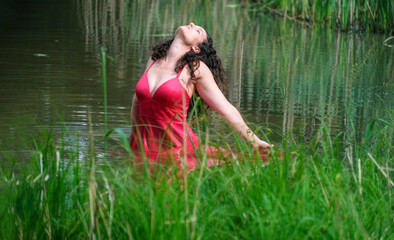 A woman in a red dress is kneeling in a tranquil pond surrounded by tall green reeds. She leans back with eyes closed, arms outstretched, embracing the peaceful, natural environment