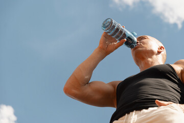 Athletes handsome sun-tanned man drinking water from bottle against sky. Concept water balance for...