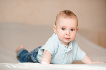 Cute smiling infant baby boy 4-5 months old crawling in bed wearing blue stylish onesie and denim pants close up. Childhood.