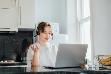 Remote work at home. Woman in headset working on Laptop at Kitchen Table in tiny flat. Student has online classes. Manager has video meeting. Customer support operator in telemarketing service