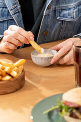 Woman deeps french fries piece into sauce at wooden table