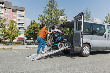 Female assistant transferring a man with disability into the vehicle by pushing his wheelchair up...