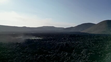An aerial drone image capturing a dramatic volcanic landscape with distant smoke rising from an active volcano. The rugged terrain, dotted with lava fields and steep hills. 4K