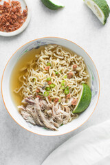 Overhead view of noodles in clear broth with shredded chicken and a lime wedge, top view of ramen noodles in chicken broth on a marble countertop