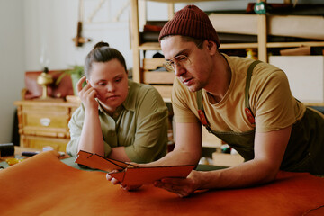 Tailor and client with down syndrome leaning on old table while examining closely leather workpieces