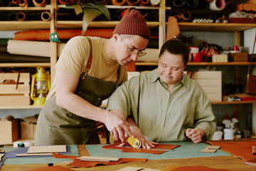 Male tailor helping his female coworker with down syndrome to cut out leather detail while they...
