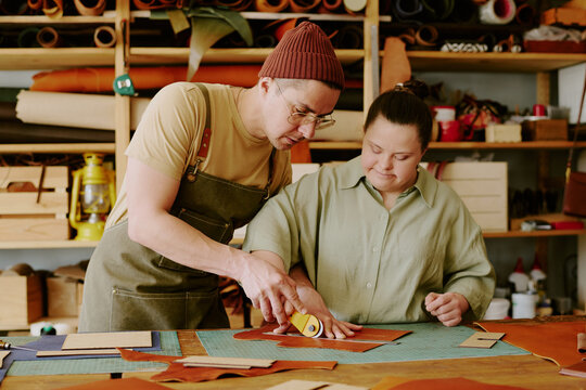 Male tailor helping his female coworker with down syndrome to cut out leather detail while they working in atelier together
