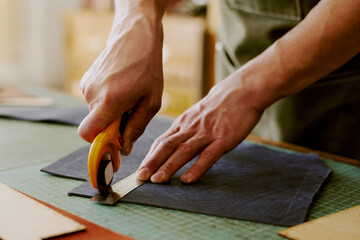 Unrecognizable male hands holding round knife and steel ruler to cut out deep blue leather workpieces