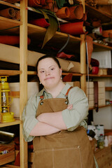 Talented female tailor with down syndrome dressed in t-shirt and chamois apron leaning on wooden shelves in atelier