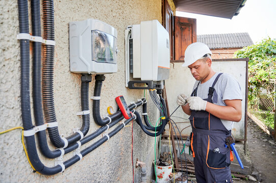 Man electrician installing solar panel system. Technician in helmet and gloves making electrical wiring inverter and electric box. Concept of alternative and renewable energy.