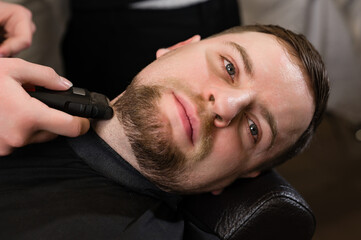 Obraz premium A barber in the salon trims the beard of a man with a electric shaver in the neck area