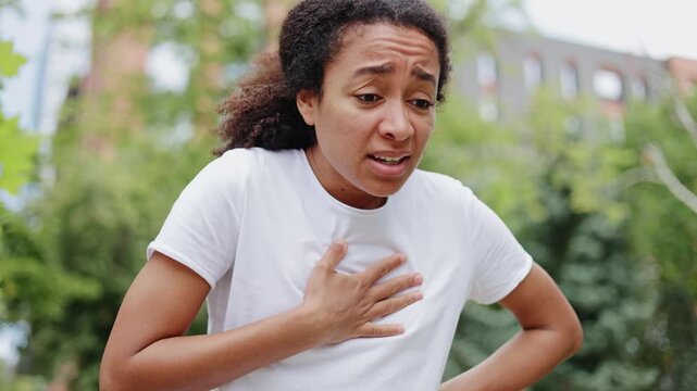 Close up of scared african american female looking around while having panic attack outdoors. Frustrated young woman touching chest and experiencing afraid emotions on background of green trees.