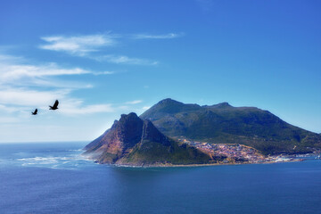 Blue sky, birds and mountain or ocean with buildings in nature or fishing village by sea. Coastal town, clouds and waves or landscape in summer for travel, vacation and outdoor for holiday by beach