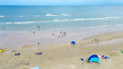 Diverse people enjoy beach activities swimming, surfing, sunbathing with umbrella, tent, camping and lounge chairs along sandy shoreline of South Beach in tropical Padre Island, Texas, aerial