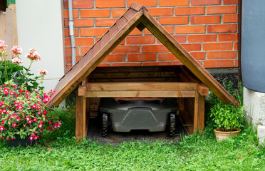 Robot lawn mower inside homemade wooden lawn mower garage outdoors in summer in home garden.