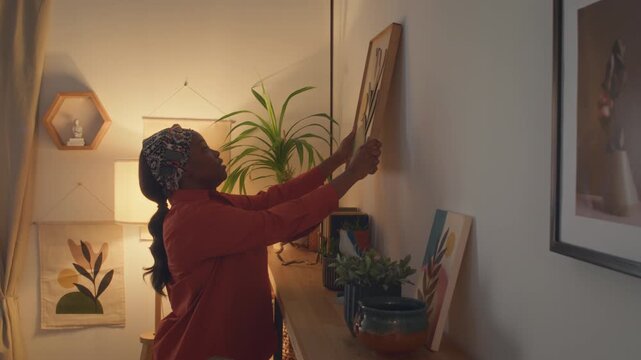 Black young woman hanging picture on wall above rack with flower pots and books standing in corner near floor lamp in living room