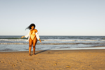 Full length photo of happy woman with afro hairstyle, overweight and obese, walking on beach in swimsuit and white shirt. Summer vacation concept of dark-skinned girl, body positive