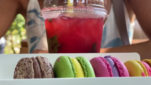 Woman drinking cold strawberry mojito cocktail with cookies at street cafe table. French macaroon sandwich cookies filled with buttercream in box.