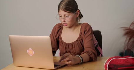 Teenage girl with glasses in distance learning sits at the table and listens attentively to the teacher, recording the lecture on her laptop. Online learning in high school. Return to school. Homework - Powered by Adobe