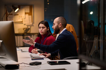 Side view portrait of bearded software developer advising young colleague and pointing at computer...
