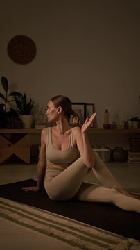 Vertical full shot of slim Caucasian lady in beige top and tights practicing yin yoga in cozy room and doing half spinal twists or ardha matsyendrasana