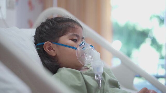 A girl is hooked up to an oxygen tank in a hospital bed. She is wearing a green gown and she is in pain.
