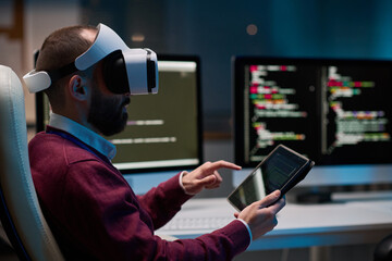 Side view portrait of bearded man wearing VR headset and using tablet working on mixed reality project in blue lights copy space