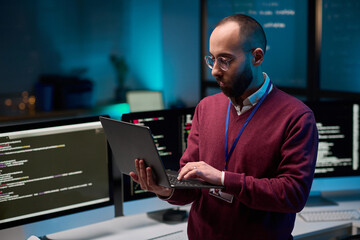 Waist up portrait of bearded adult man holding laptop while standing in cybersecurity office with code lines on screens copy space