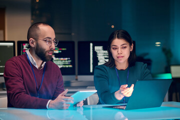 Portrait of young woman as female IT security expert using laptop with male colleague in blue neon...