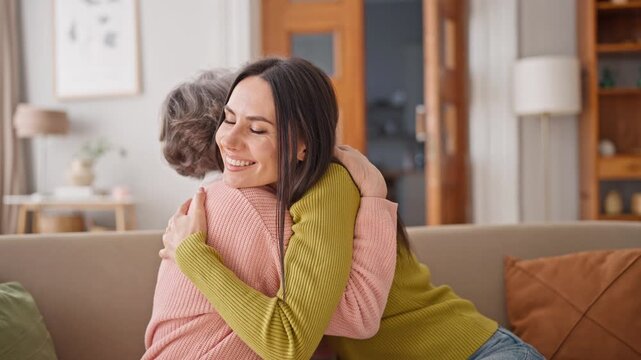 Happy Adult Caucasian mother enjoying great free time with her beautiful daughter. Relatives hugging each other while sitting on sofa in cosy room. Warm moment. Family smiling to each other with love.