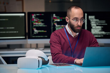 Portrait of bearded adult man wearing glasses and using laptop at desk in IT development office ,...