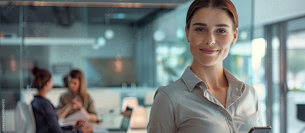 Poster a portrait showing a caucasian lady smiling holding a smartphone in a modern office with a colleague