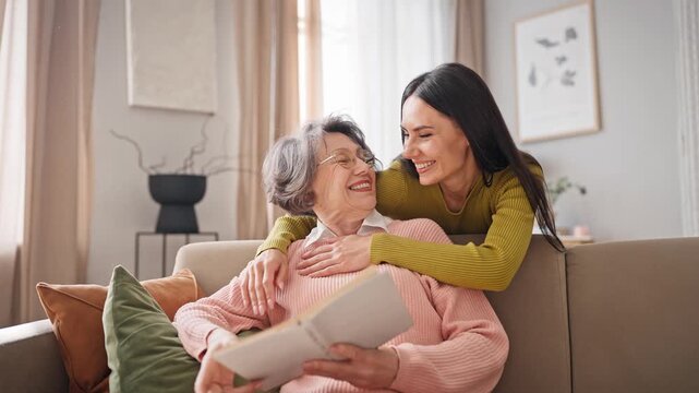 Elderly Caucasian woman with grey hair wearing glasses while reading book. Charming daughter walking up behind mature mother and hugging her. Older mom widely smiling while looking at her relative.