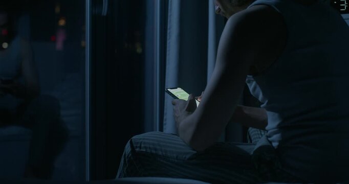 A man uses a mobile phone while sitting on the bed at night. Close-up of his hands, an unrecognizable person. Dark room, home interior