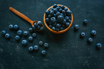 Wooden bowl filled with fresh blueberries on a dark textured background. A wooden spoon lies beside it, with scattered berries creating a rustic and elegant presentation