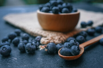 Wooden bowl filled with fresh blueberries and a wooden spoon with a few berries on a dark surface. The rustic presentation highlights the natural beauty and vibrant color of the fruit
