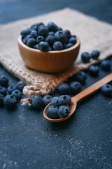 Wooden bowl filled with fresh blueberries and a wooden spoon with a few berries on a dark surface. The rustic presentation highlights the natural beauty and vibrant color of the fruit