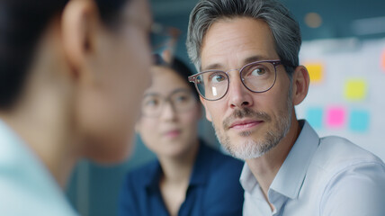A middle-aged man with glasses engages in discussion with colleagues in a modern office. Blurred sticky notes in the background emphasize brainstorming and collaboration.