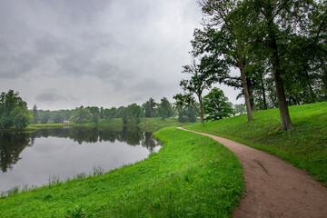 A tranquil pathway winds alongside a calm lake, surrounded by vibrant greenery and tall trees, inviting peaceful reflections on a cloudy day.