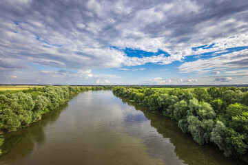 A peaceful river meanders through vibrant green foliage, reflecting the dynamic clouds above in a...