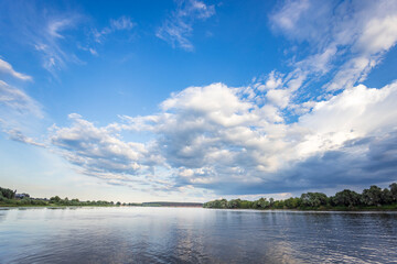 A wide, calm river stretches out under a bright blue sky, dotted with fluffy white clouds. Green trees line the riverbanks, creating a tranquil and peaceful scene.