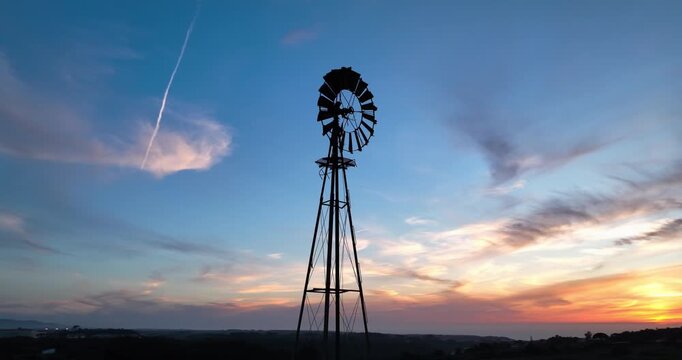 A vintage metal windmill in rural Portugal powers a farm water pump in the dry outback. A drone shot captures blades rotating against a sunset backdrop, showcasing the timeless beauty of this scene.