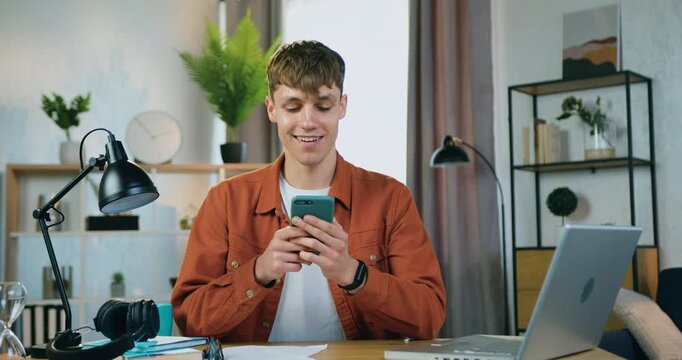 Trendy young man sitting at the table at home and using his smartphone to write message to his mates. Leisure concept
