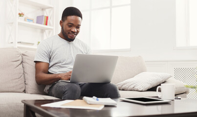 Young happy black man chatting online on laptop with friend at home. Casual guy sitting on beige couch in light livingroom, copy space